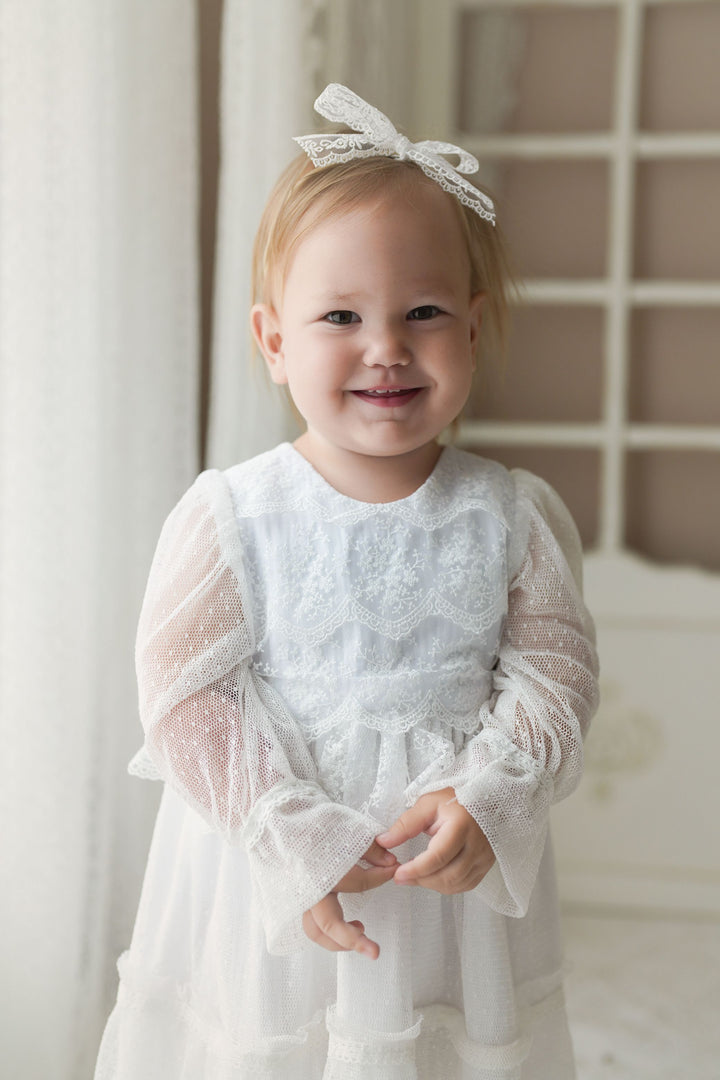 A young girl in a white lace dress with a bow on her head is smiling and posing for a photograph.
