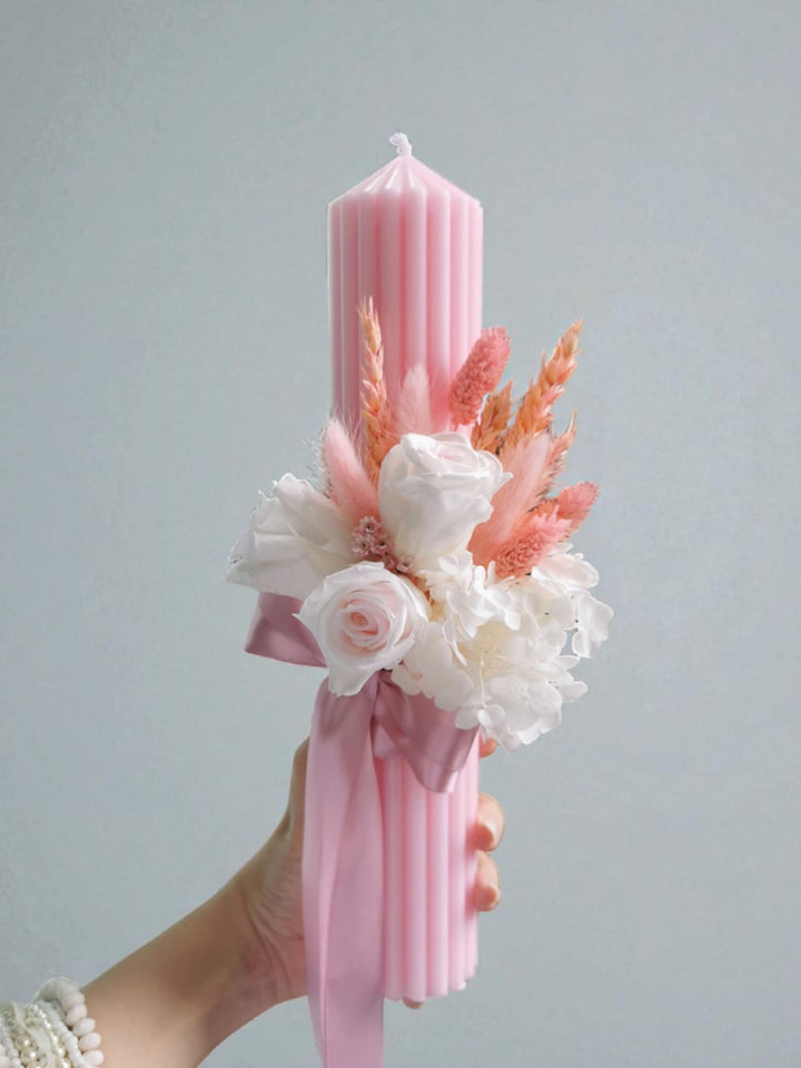 A hand holding a pink candle with a floral arrangement on top.
