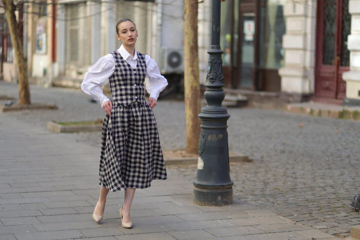 A woman in a white and black checkered dress stands on a cobblestone street, with a lamppost and buildings in the background.