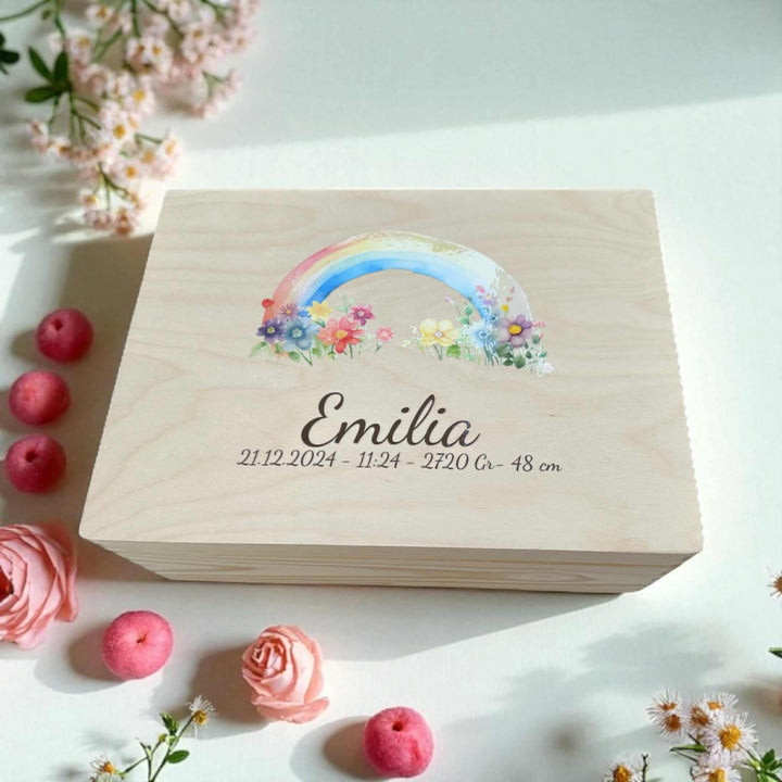 A wooden box with a colorful rainbow graphic and the name "Emilia" printed on it, surrounded by pink flowers and small red apples.