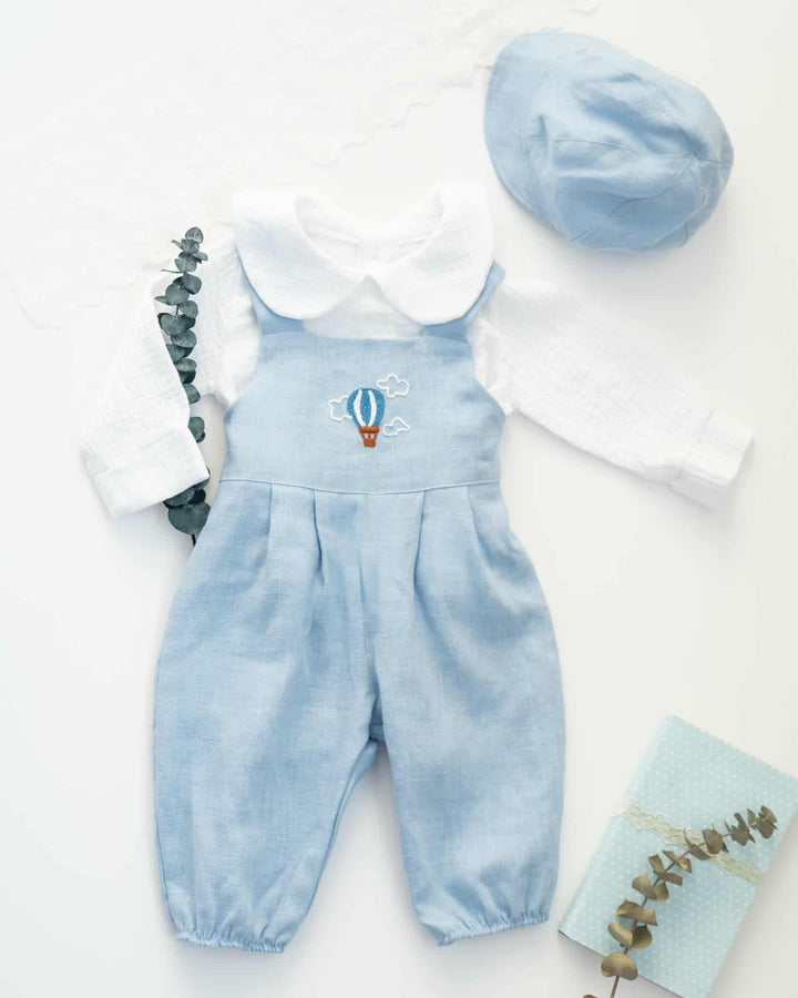 A light blue onesie with a white collar and a hot air balloon design, a white long-sleeved shirt, and a light blue cap are arranged on a white background.