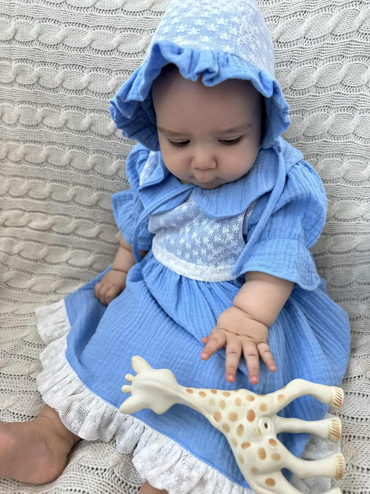 A baby girl in a blue dress and bonnet is sitting on a white blanket, holding a white giraffe toy.
