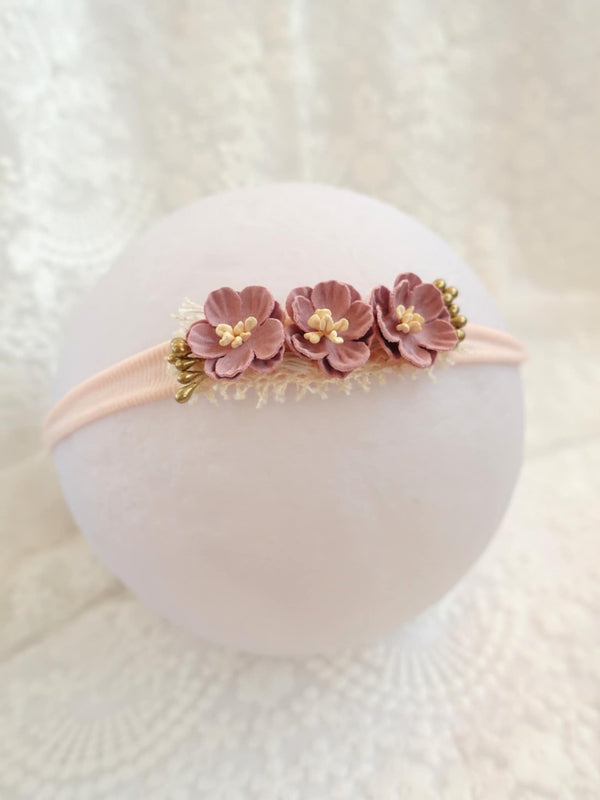 A delicate pink flower headband with three petals and gold accents rests on a white surface.
