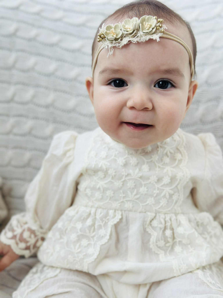 A young baby girl wearing a white lace dress and a flower headband sits on a white couch, looking directly at the camera with a smile.