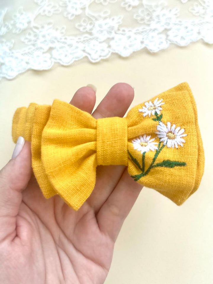 A hand is holding a yellow bow with white daisies embroidered on it.
