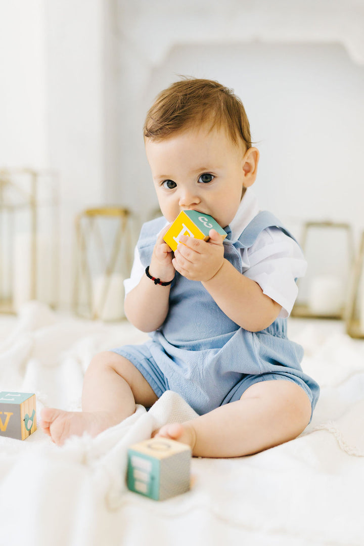 A baby boy sitting on a white blanket, holding a yellow and green block in his mouth.