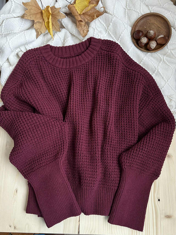 a burgundy-colored knitted sweater laid out on a wooden surface, with a bowl of chestnuts and autumn leaves in the background.