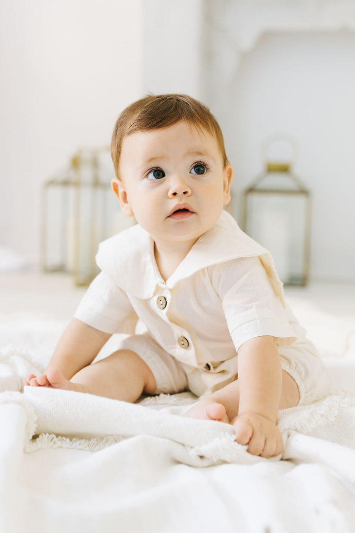 A baby in a white outfit sits on a white blanket, looking up with a curious expression.