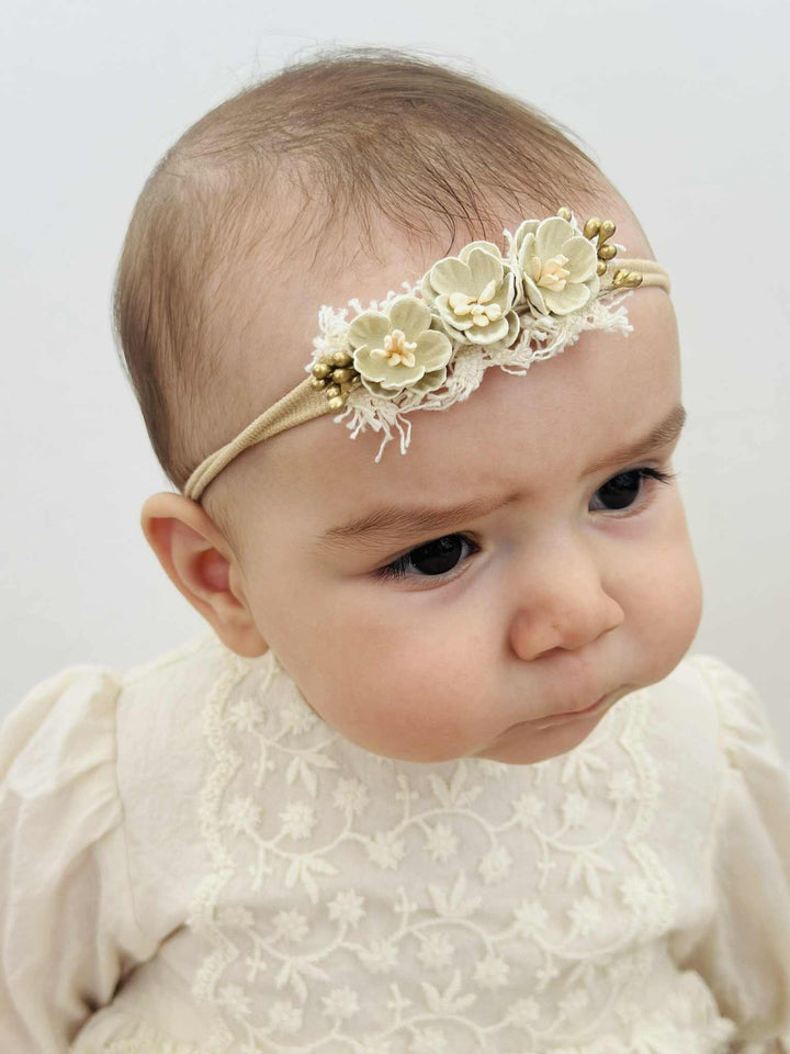 A close-up portrait of a baby girl wearing a white lace dress and a flower headband.