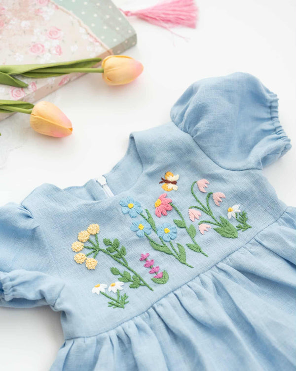 A light blue dress with colorful floral embroidery, including flowers and leaves, is displayed on a white surface alongside a pink tassel and a small floral box.