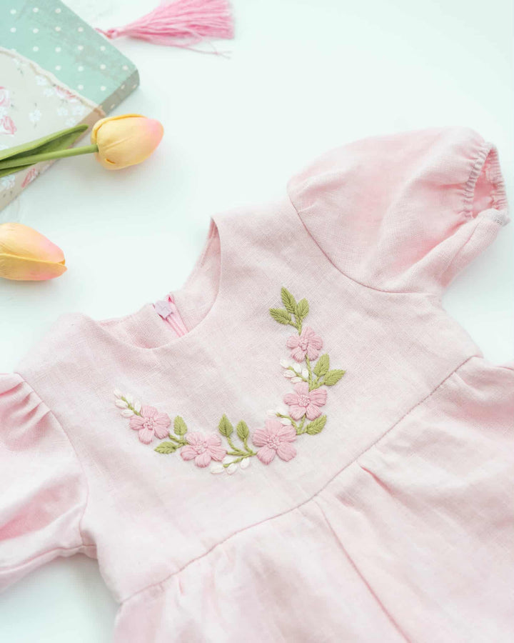 A pink dress with a floral embroidered design on the front, placed on a white surface alongside a green box and two yellow tulips.