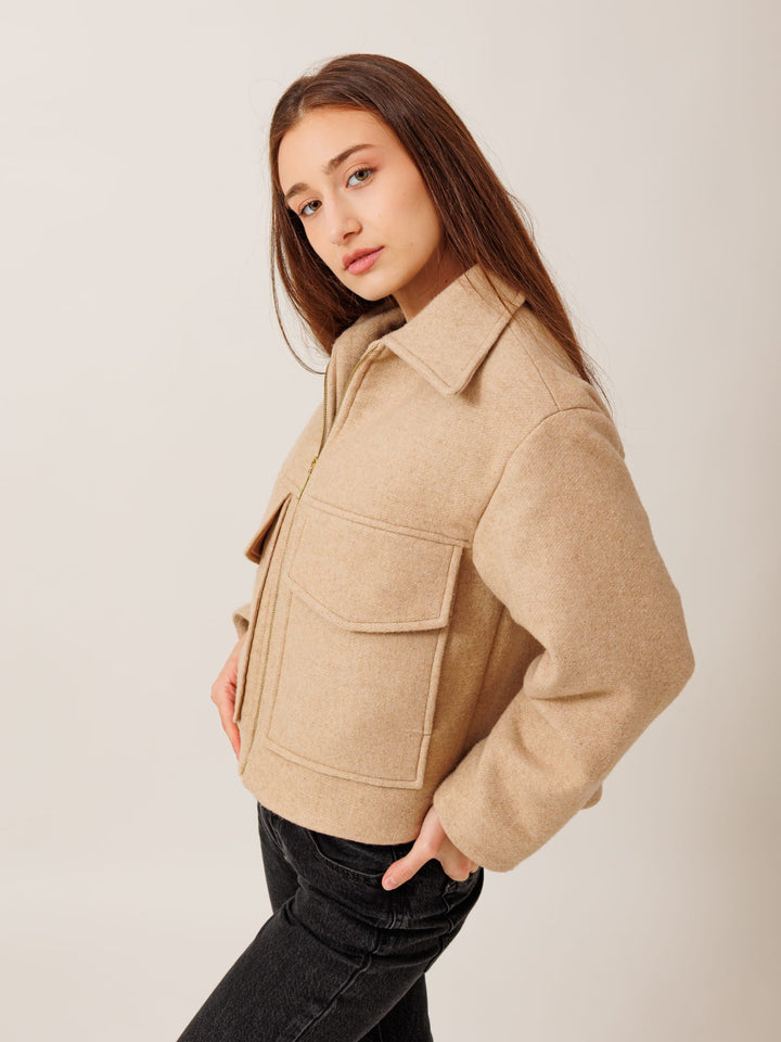 A young woman with long brown hair wearing a beige jacket and black pants stands against a plain background, looking directly at the camera with a serious expression.
