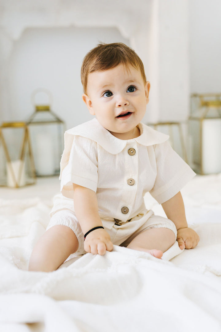 A smiling baby boy wearing a white outfit sits on a white blanket, surrounded by gold lanterns in the background.
