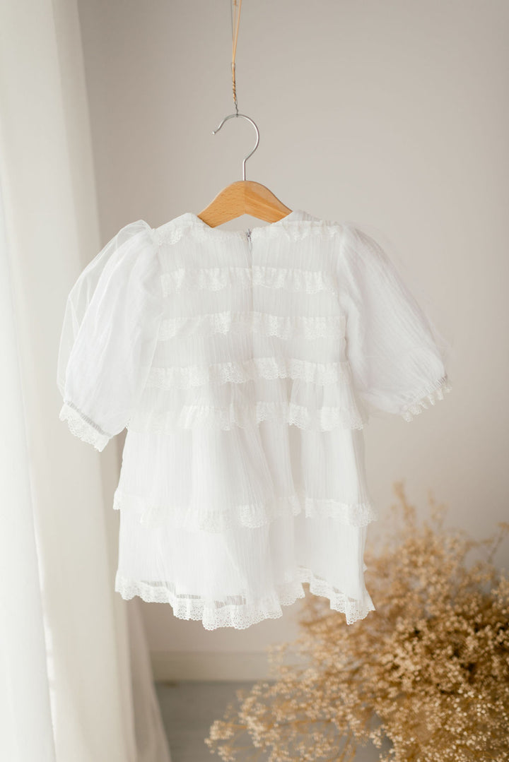 A white, ruffled dress with lace trim hangs on a wooden hanger against a white wall.