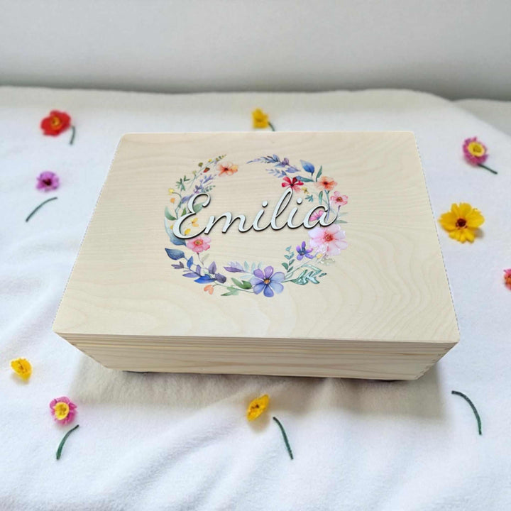 A wooden box with the name "Emilia" printed on it, surrounded by colorful flowers on a white surface.