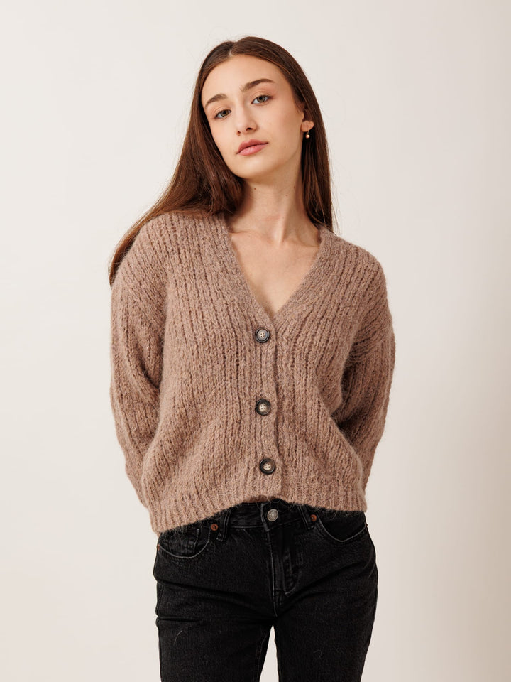 A young woman with long brown hair wearing a brown cardigan and black pants stands against a plain white background.