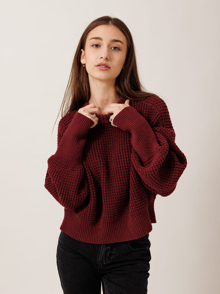 A young woman with long brown hair wearing a burgundy knitted sweater stands against a plain background, looking directly at the camera with a neutral expression.