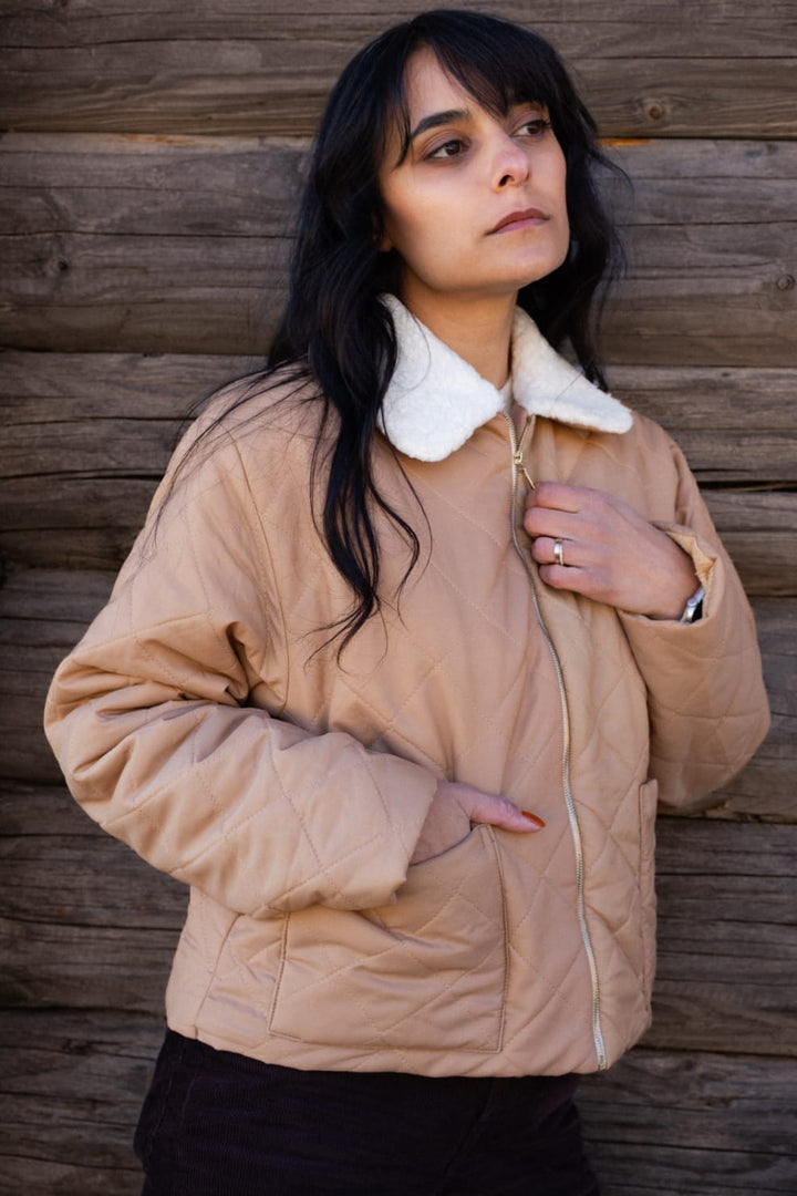 A woman wearing a beige jacket with a white collar stands in front of a wooden wall, looking off to the side with a serious expression.