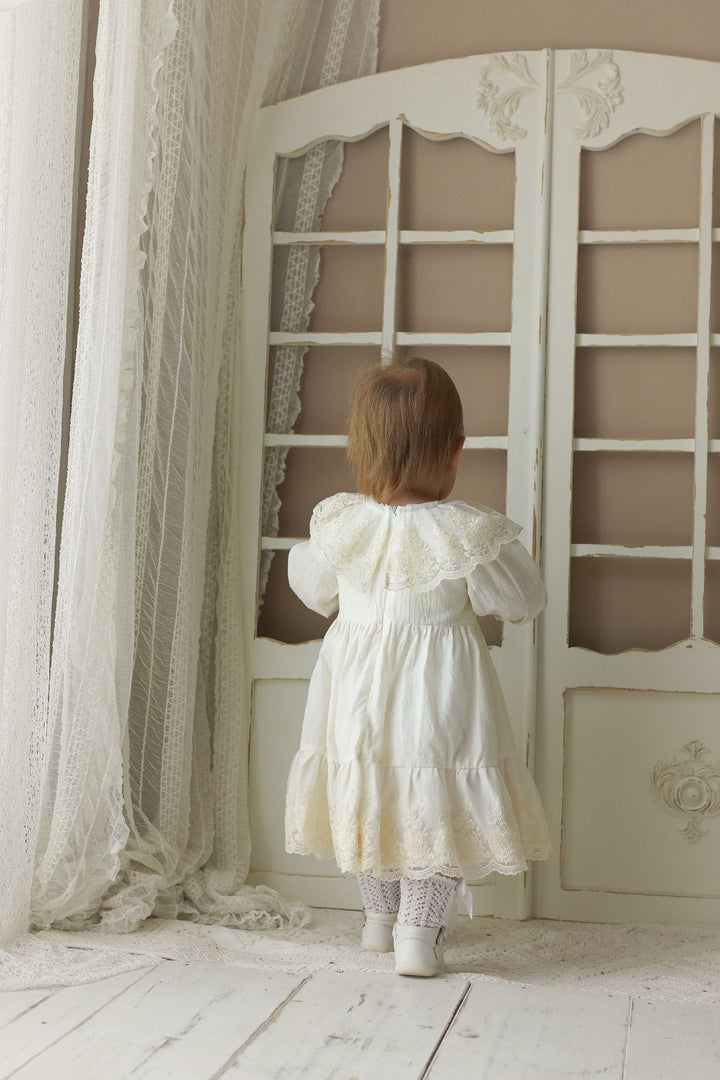 A young child in a white lace dress stands in front of a white lace curtain, looking out of a window.