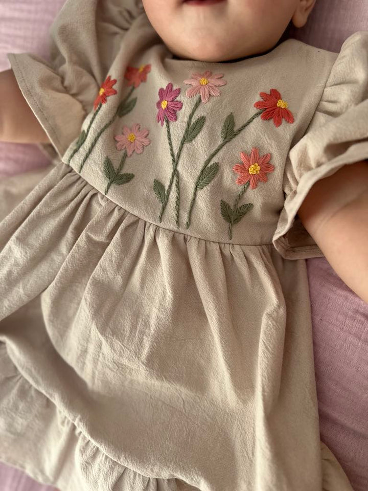 A baby wearing a beige dress with colorful floral embroidery on the front.