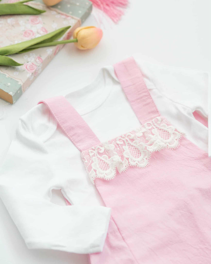A pink and white baby outfit with lace trim, including a white shirt and pink suspenders, is displayed on a white surface alongside a book and a pink flower.