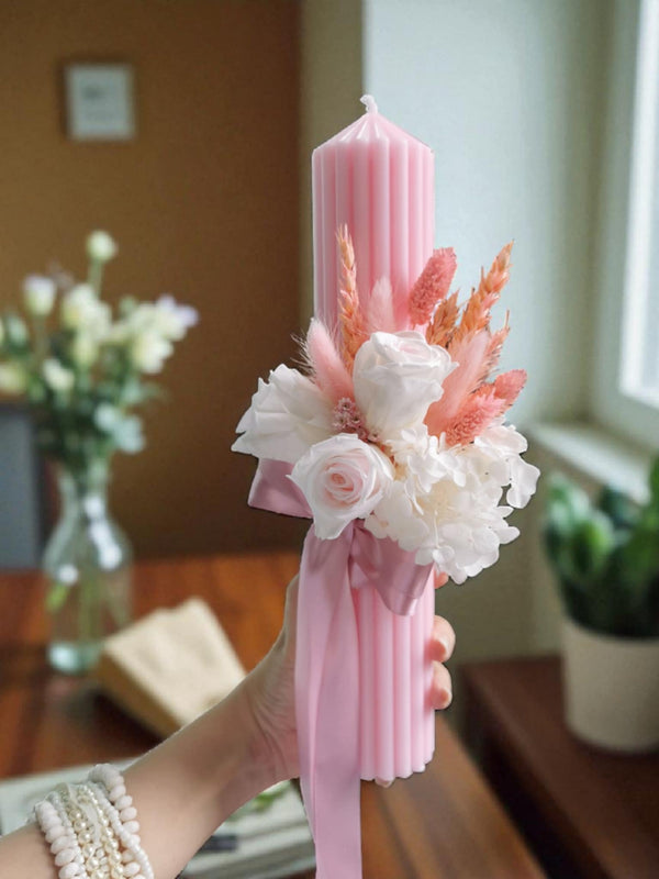 A hand holding a pink candle with a floral arrangement on top, set against a backdrop of a vase of flowers and a window.