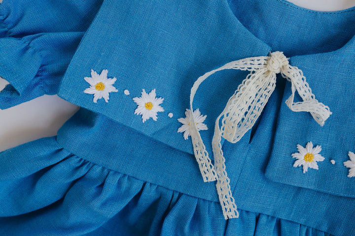 A close-up view of a blue dress with white daisy embroidered flowers and a white lace bow.