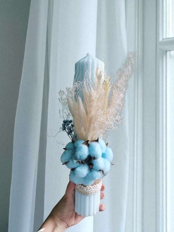 A hand holding a white vase filled with blue cotton balls and dried flowers, placed in front of a white curtain.