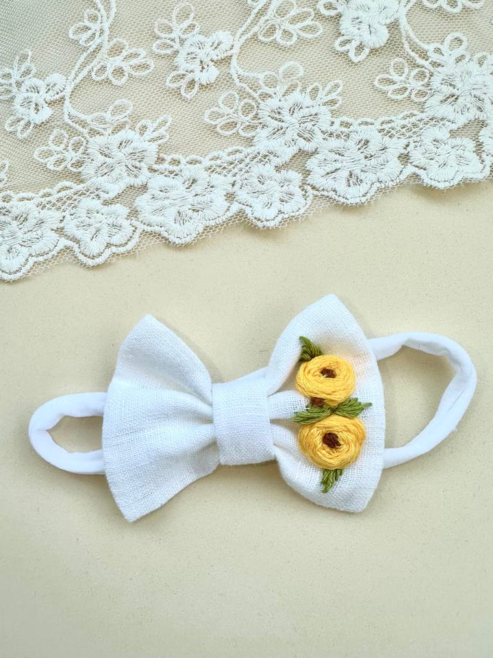 A white bow with yellow flowers is placed on a beige background, with a white lace doily visible in the top left corner.