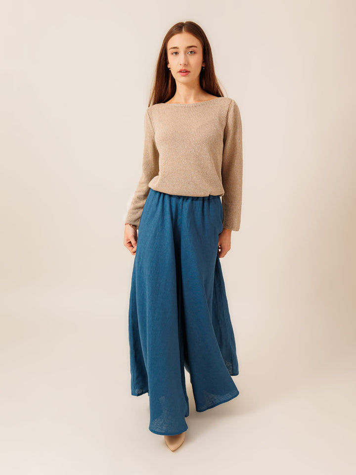A woman wearing a beige sweater and a long blue skirt stands against a plain background.