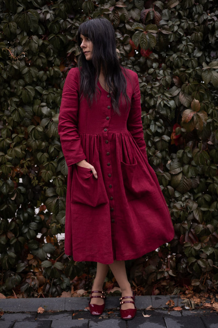A woman in a long red dress with button-up sleeves stands in front of a lush green hedge.