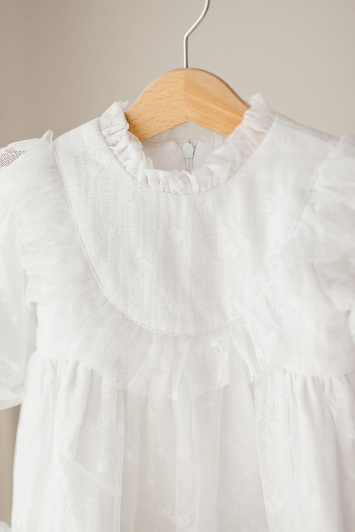 A white, ruffled dress with a lace collar hangs on a wooden hanger.