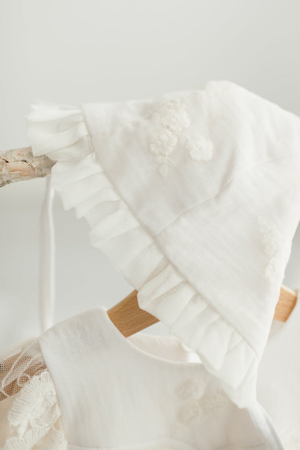 A white lace bonnet with ruffled edges is hanging on a wooden hanger.