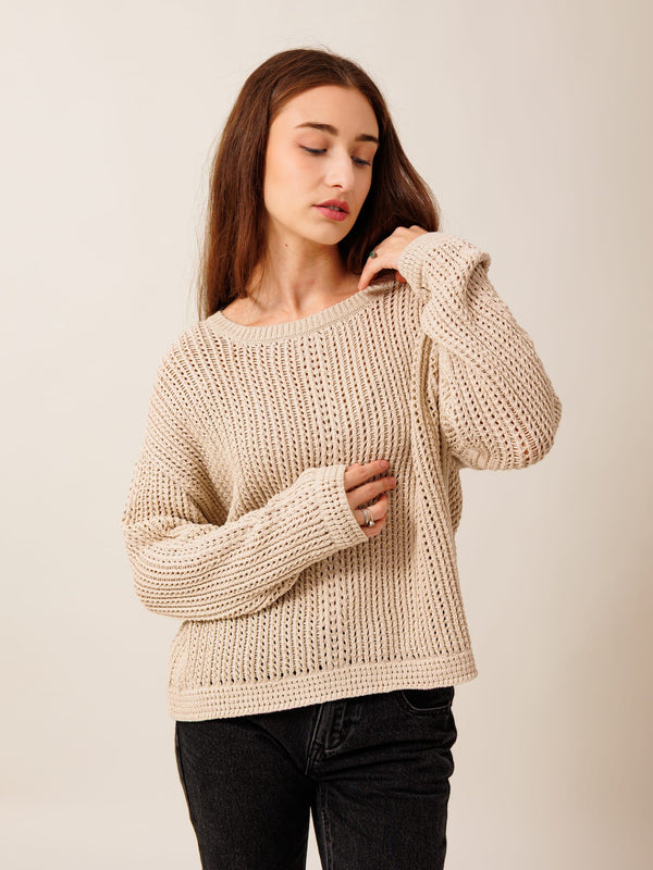 A young woman with long brown hair wearing a beige knitted sweater stands against a plain background, looking down at her hands.
