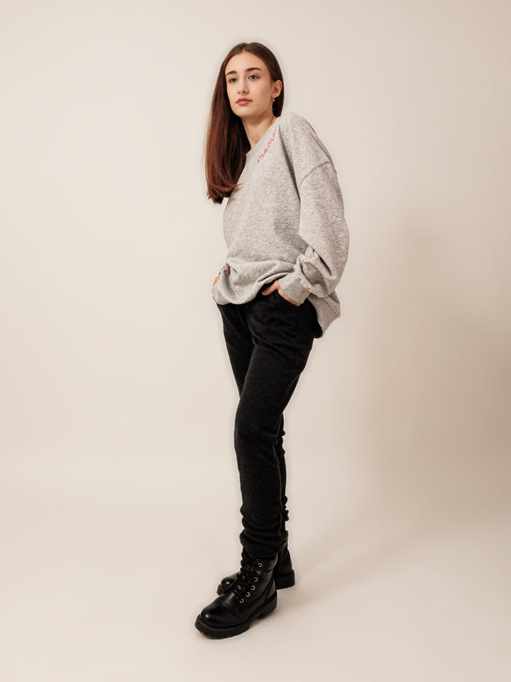 A young woman with long brown hair stands against a plain white background, wearing a gray sweatshirt, black pants, and black boots.
