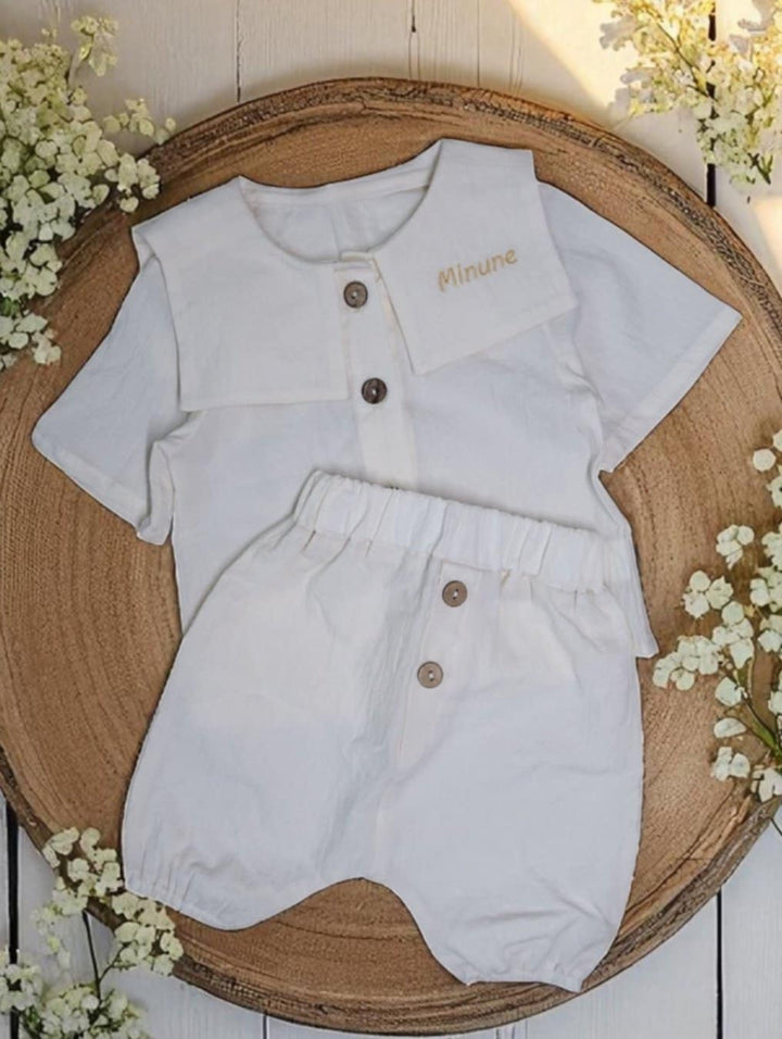 A white baby outfit with a collar and buttons, along with matching white shorts, is displayed on a wooden tray. The outfit is placed on a wooden surface, and there are white flowers in the background.