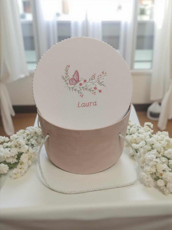 A round white box with a pink handle and a butterfly design, sitting on a white tablecloth. The box has a name written on it.