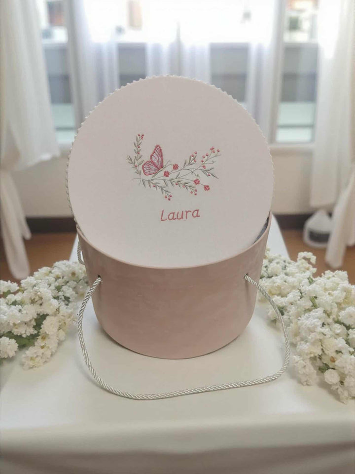 A round white box with a pink handle and a butterfly design, sitting on a white tablecloth. The box has a name written on it.