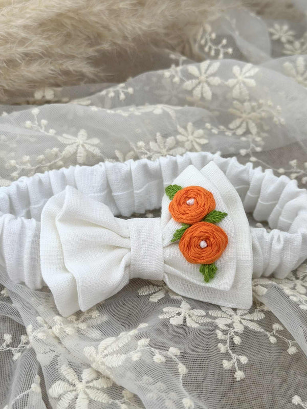 A white garter belt with a bow made of orange yarn and green leaves, resting on a white lace background.