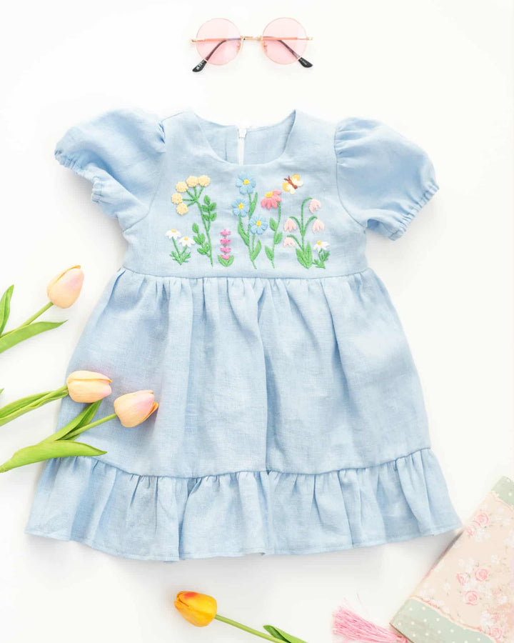 a light blue dress with a floral embroidered design, accompanied by pink sunglasses and a bouquet of pink and yellow tulips.
