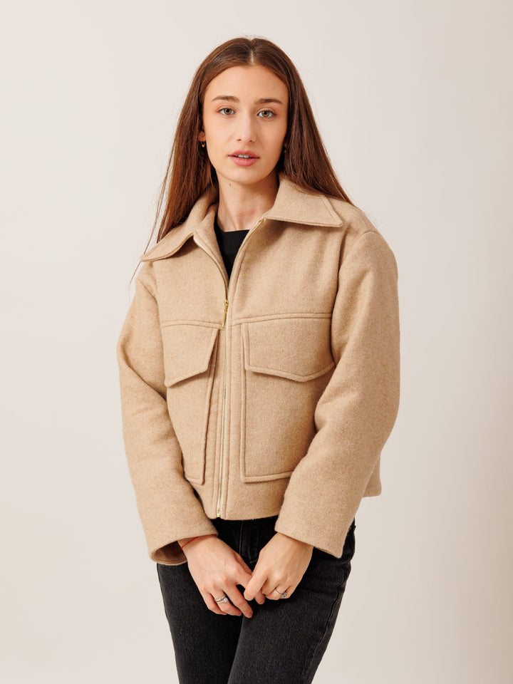 A young woman with long brown hair wearing a beige jacket with a zipper and two pockets stands against a plain background.