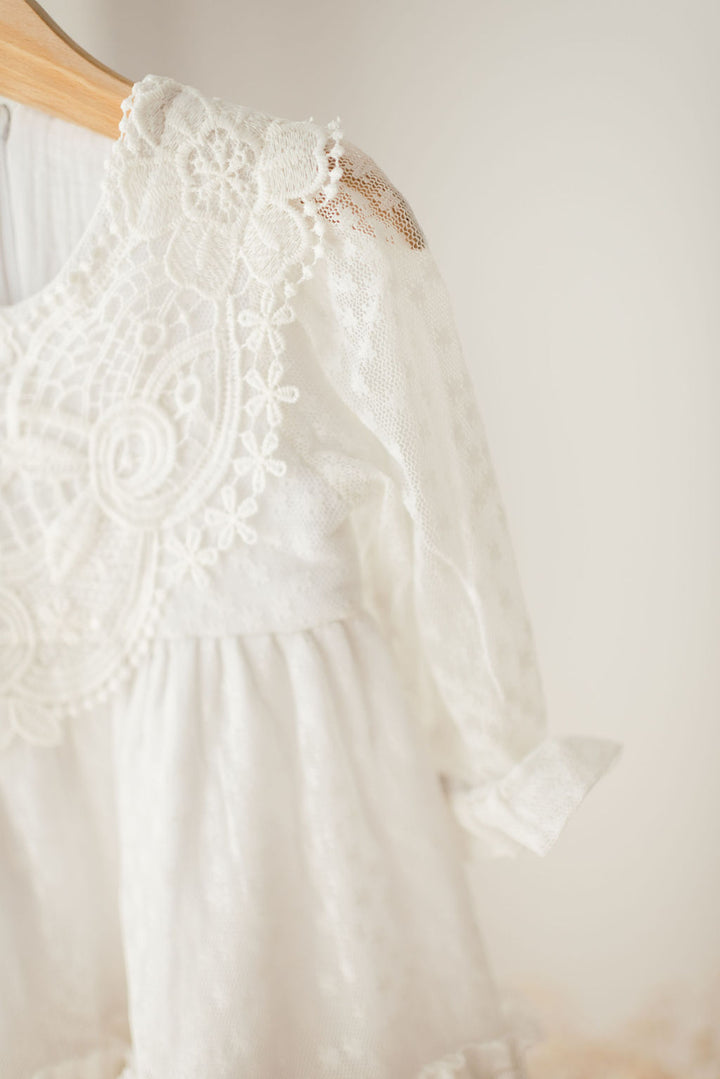A close-up view of a white lace dress with intricate floral patterns, hanging on a wooden hanger against a plain background.