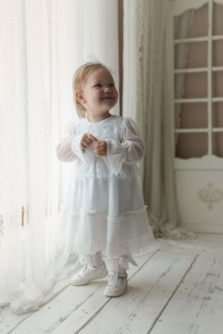 A young girl in a white lace dress stands in a bright, white room, smiling and looking up.