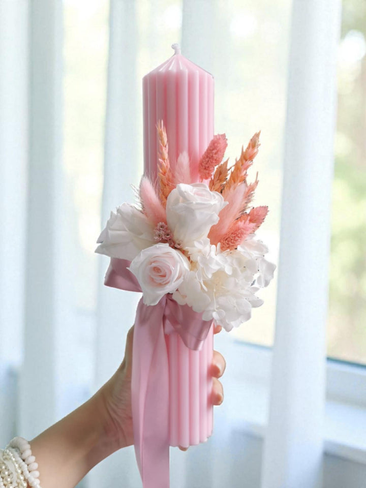 A hand holding a pink candle with a floral arrangement on it, placed in front of a window.