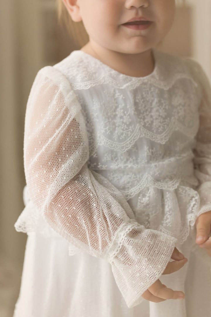 A young girl in a white lace dress with a ruffled collar and sleeves stands in a room.