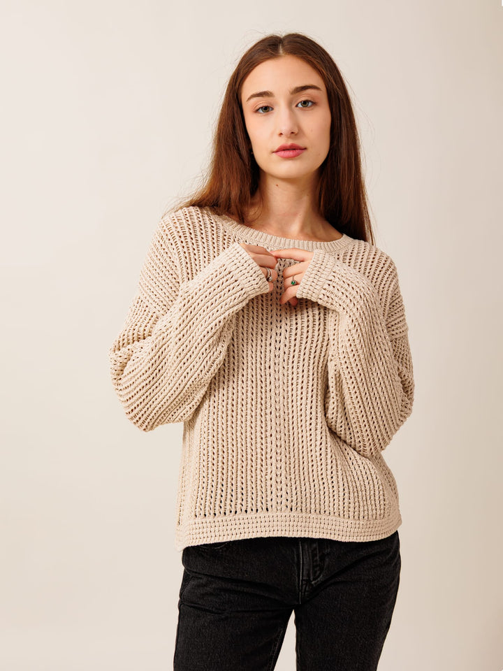 A young woman with long brown hair wearing a beige knitted sweater and black pants stands against a plain background, looking directly at the camera with a serious expression.