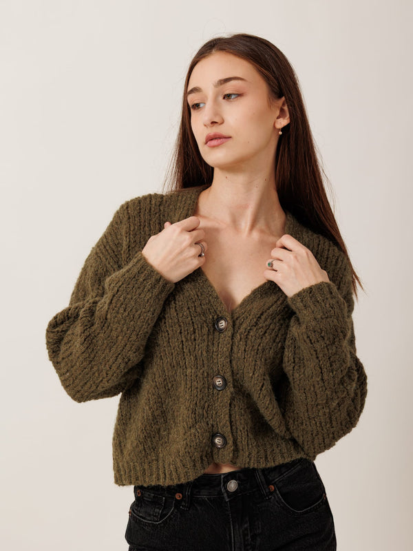 A young woman with long brown hair wearing a green cardigan and black pants stands against a plain white background, looking to her left with a thoughtful expression.