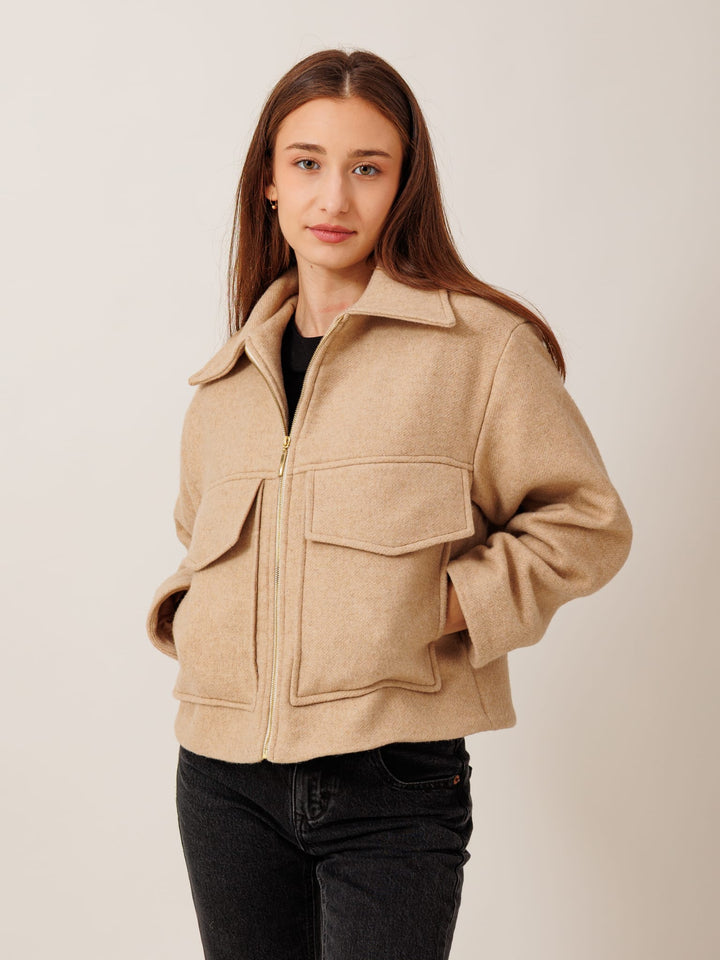 A young woman with long brown hair wearing a beige jacket with a zipper and pockets, standing against a plain background.