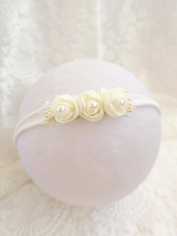 A white headband adorned with three white roses and pearls rests on a white surface.
