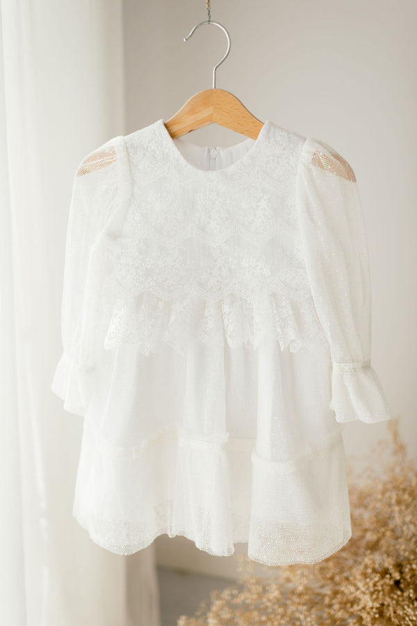 A white lace dress with ruffled sleeves and a ruffled hem hangs on a wooden hanger against a white background.